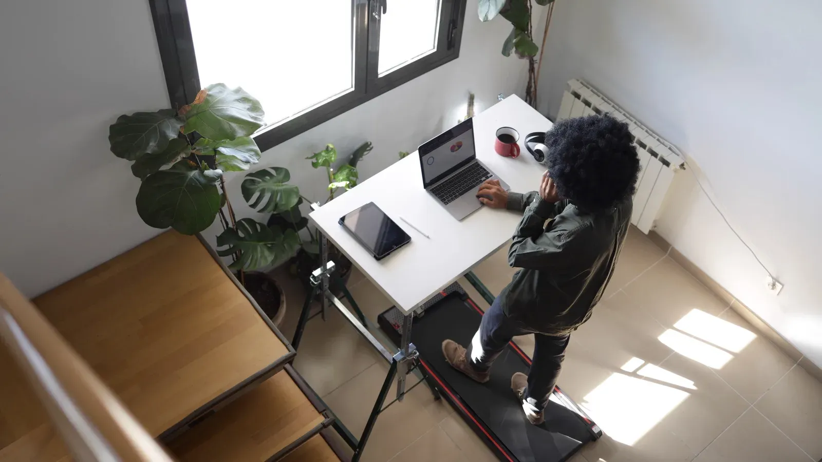 A person walking on a compact treadmill under a standing desk while working on a laptop